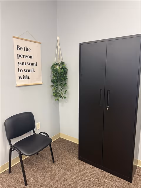 A small office corner with a black cabinet, a chair, hanging plant, and inspirational poster.