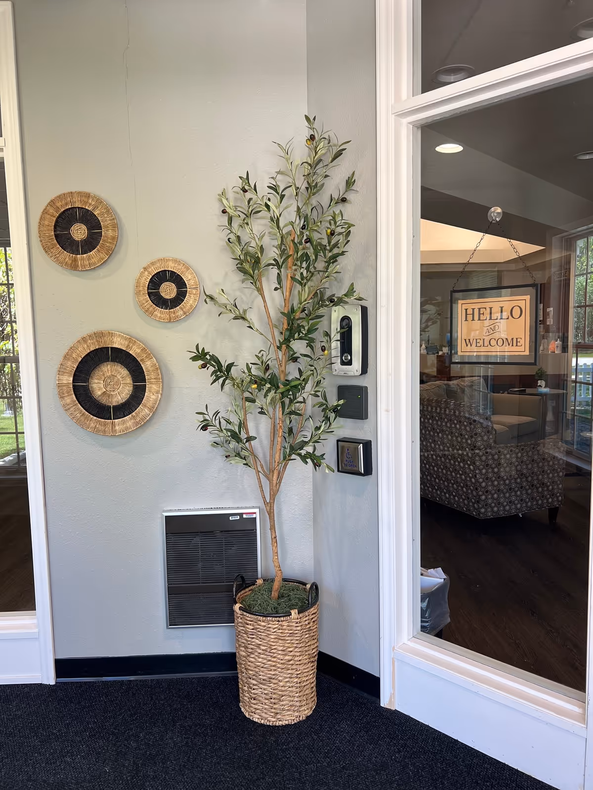 Indoor lobby with a potted olive tree, woven wall decor, and Hello and Welcome sign.