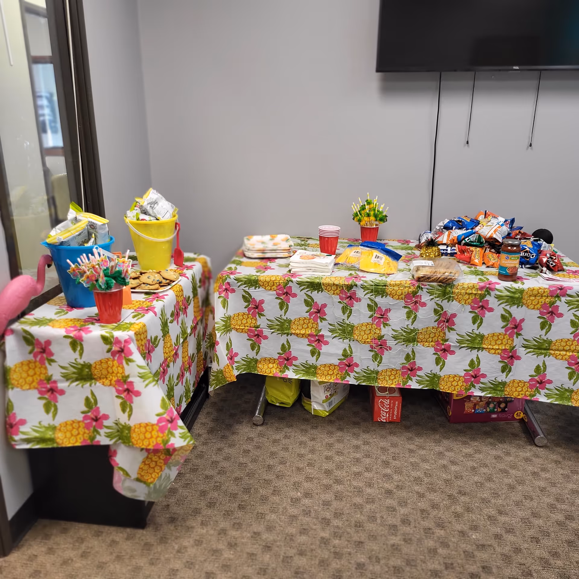 Two tables with a pineapple pattern tablecloth display snacks, candy, cups, and decorations indoors.