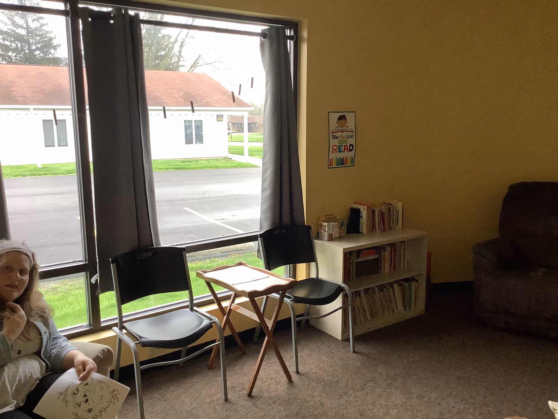 Cozy senior living lounge with large windows, chairs, a bookshelf, and a resident seated left.