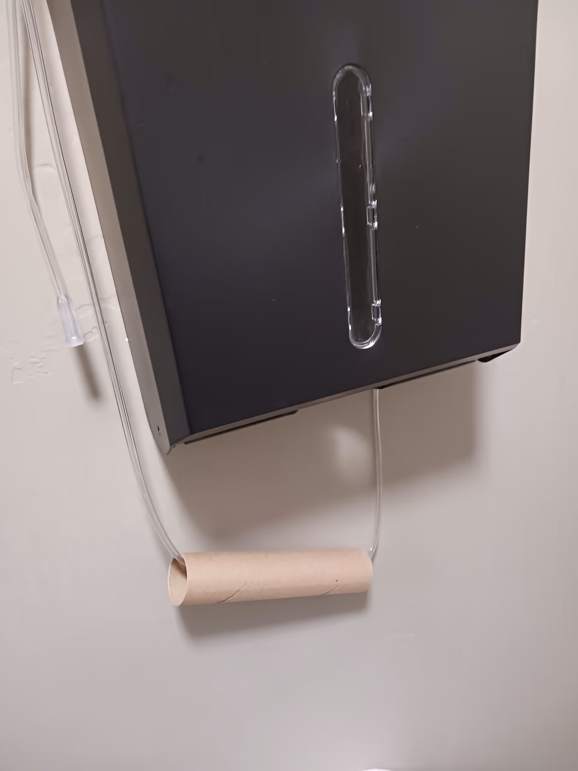 Black wall-mounted dispenser with a clear window on a white bathroom wall.