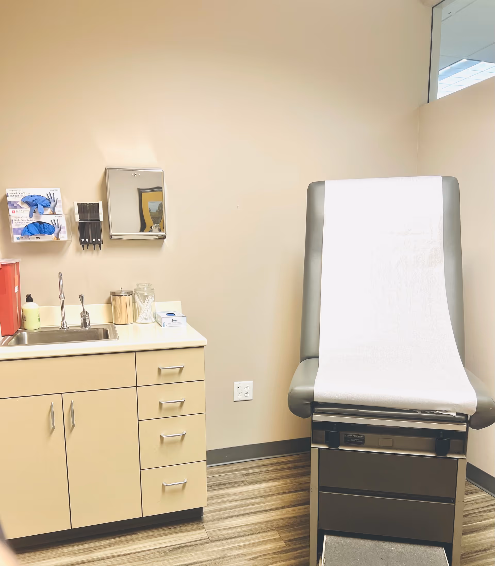 A clinical examination room with sink, cabinets, and a paper-covered exam chair.