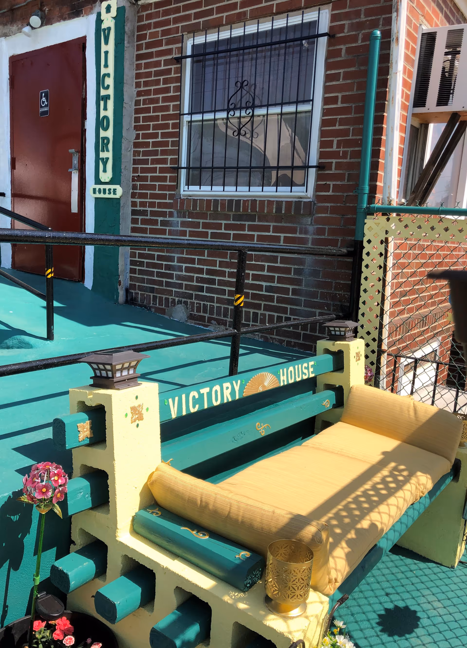 Outdoor seating area with turquoise bench beside Victory House entrance at brick building.
