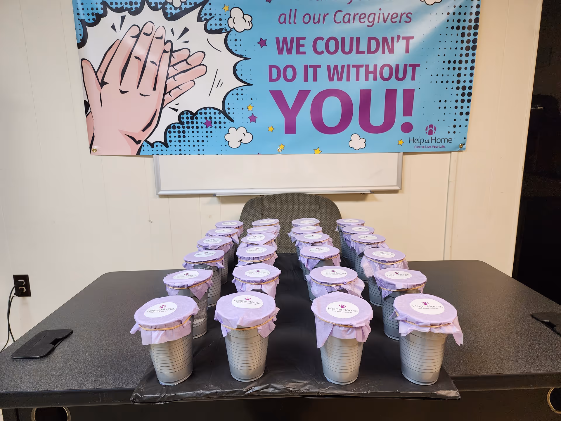 Row of small cups with purple covers on a table, banner for Help at Home behind.