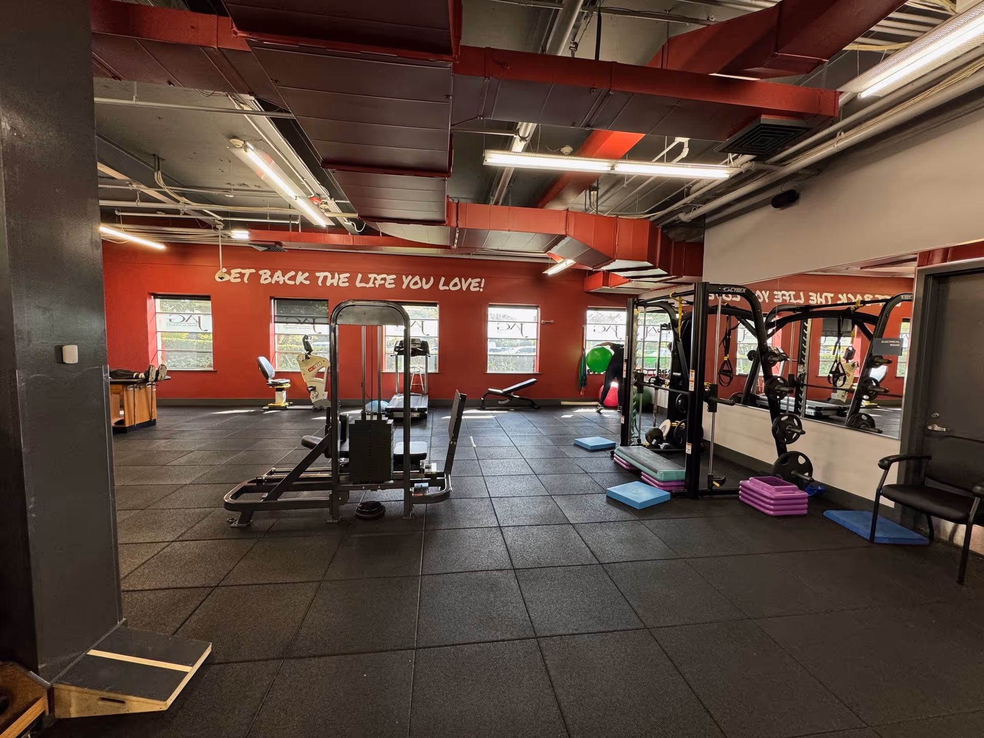 Red-walled gym area with exercise machines and mirrors inside a rehab facility.