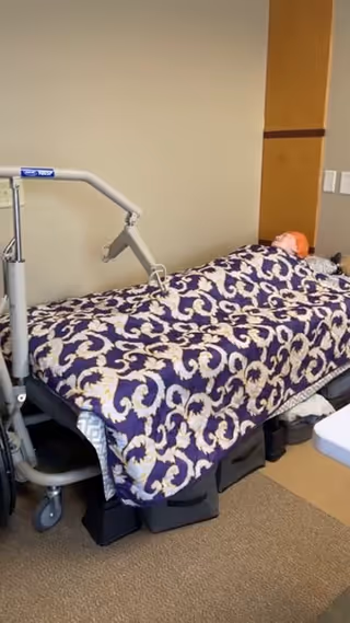 Facility bedroom with an adjustable bed, purple patterned blanket, and safety equipment.