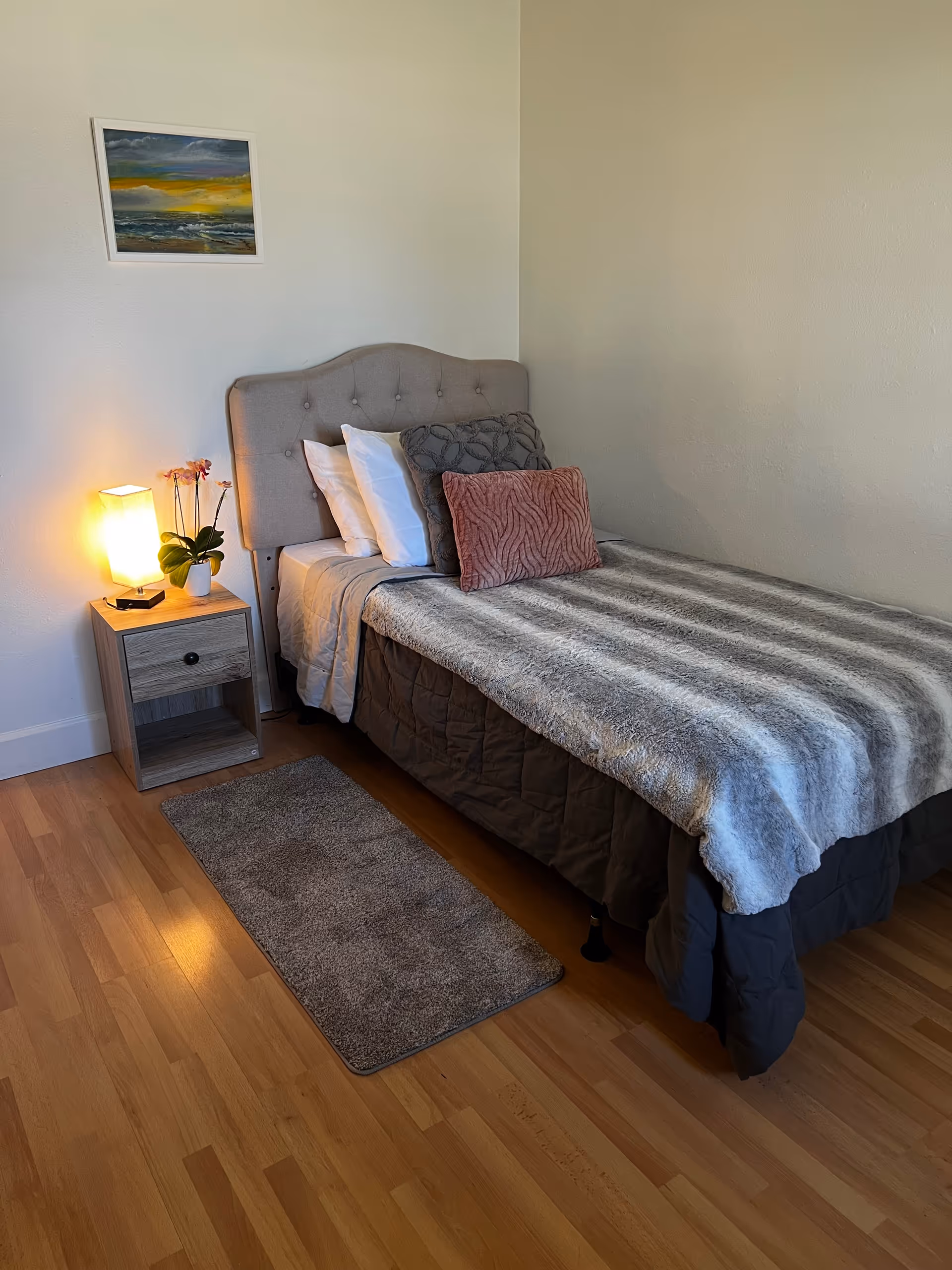 A neatly made bedroom with a single bed, nightstand, lamp, rug, and wall art.
