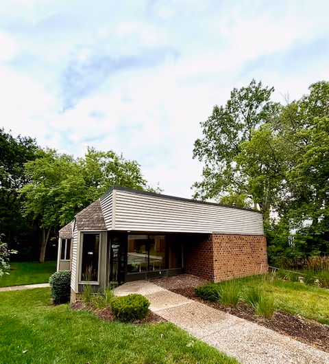 A small brick and siding building with a glass entrance, surrounded by green lawn and trees.