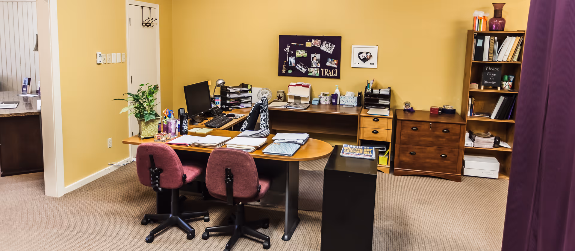 A tidy office area with a curved desk, chairs, computer, and filing shelves.
