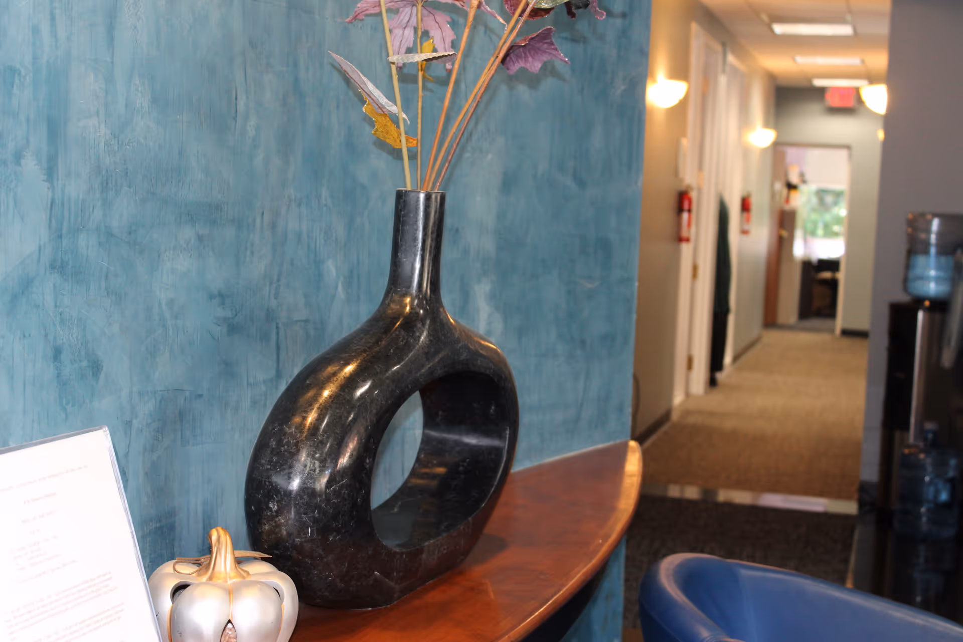 Interior hallway of a senior facility with teal wall and black vase on a curved table.