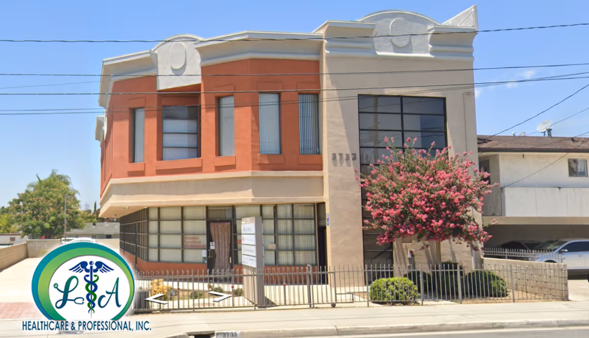 Two-story building with beige and reddish facade, large storefront windows, and blooming tree.