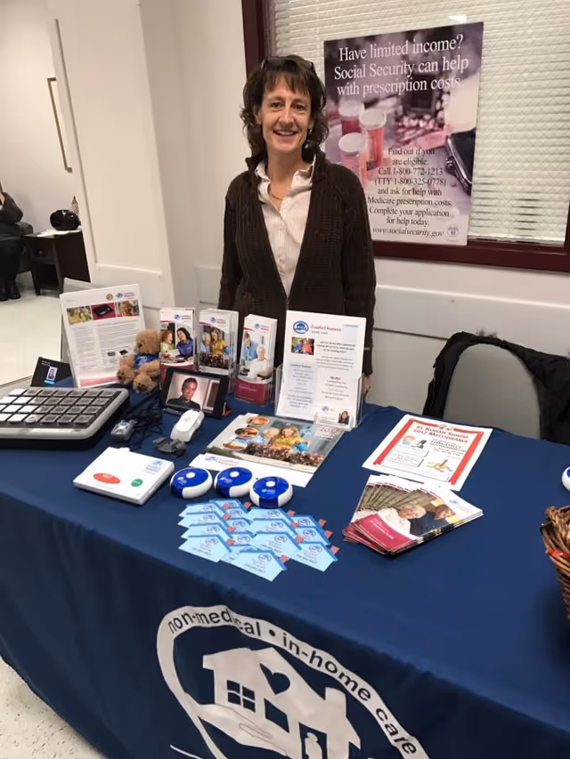 Smiling woman stands behind a table of brochures at an information booth inside a facility.
