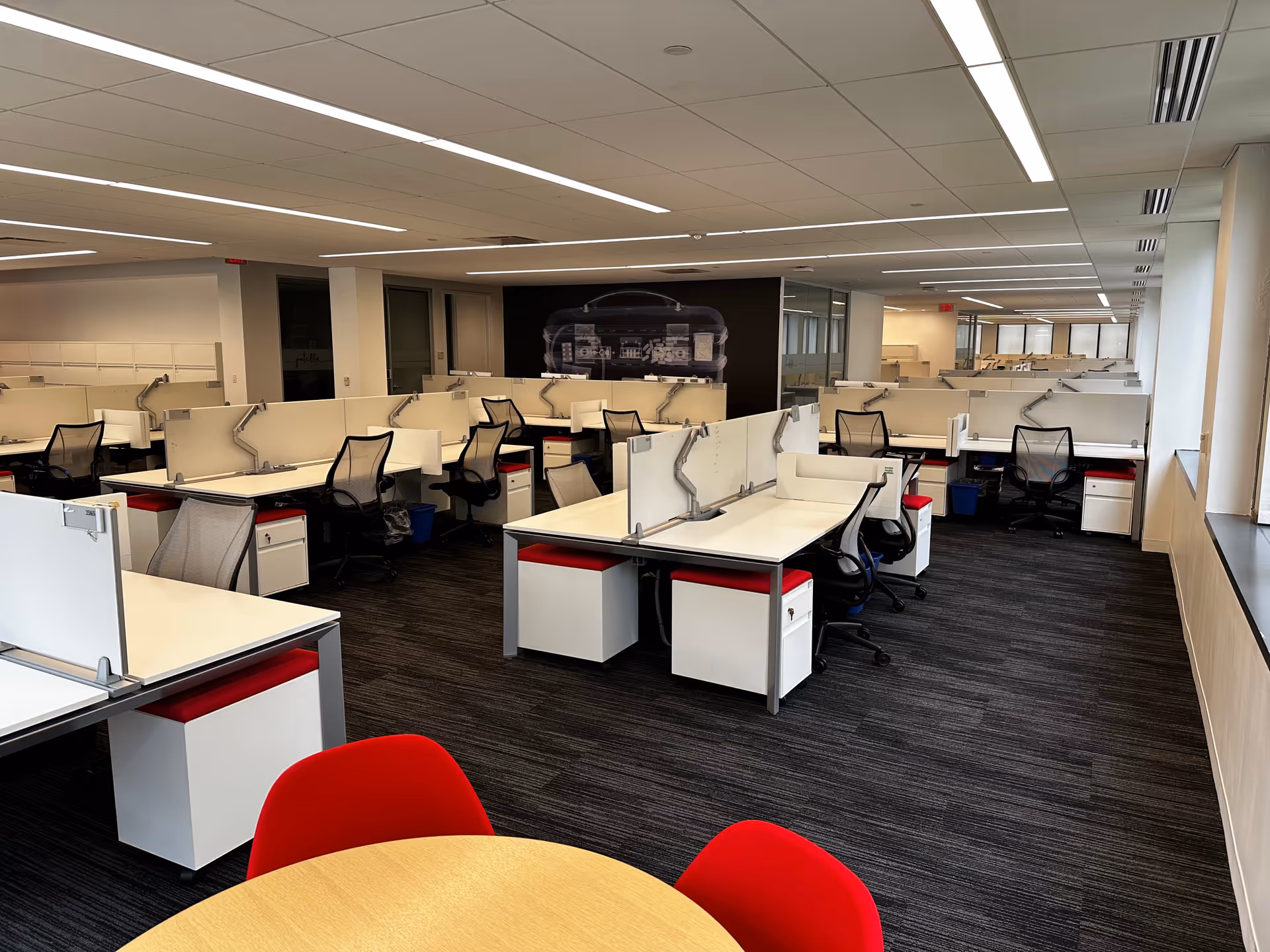 Open-plan office with workstations, cubicles, and red-accent chairs under bright overhead lighting.