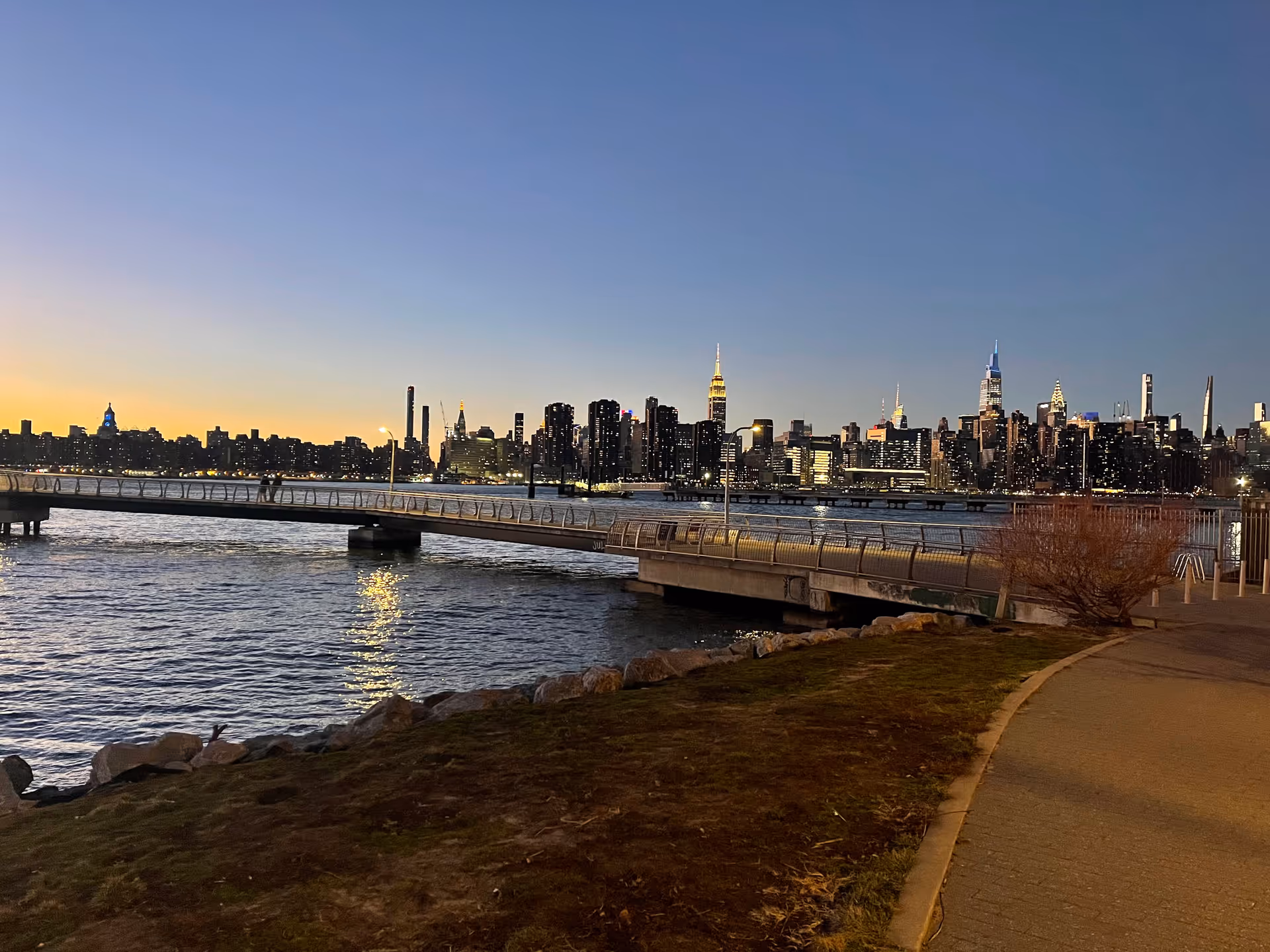 Sunset over a city skyline viewed from a riverside walkway with railings.