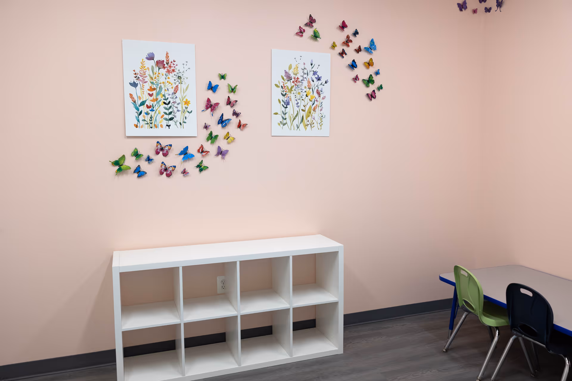 Soft pink wall with butterfly wall art, cubby shelf, and small table with colorful chairs.