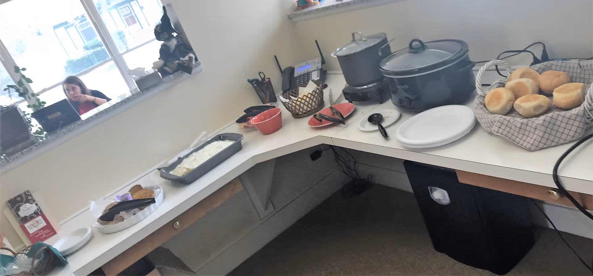 A bright facility kitchen counter with a slow cooker, bread rolls, and prep items.