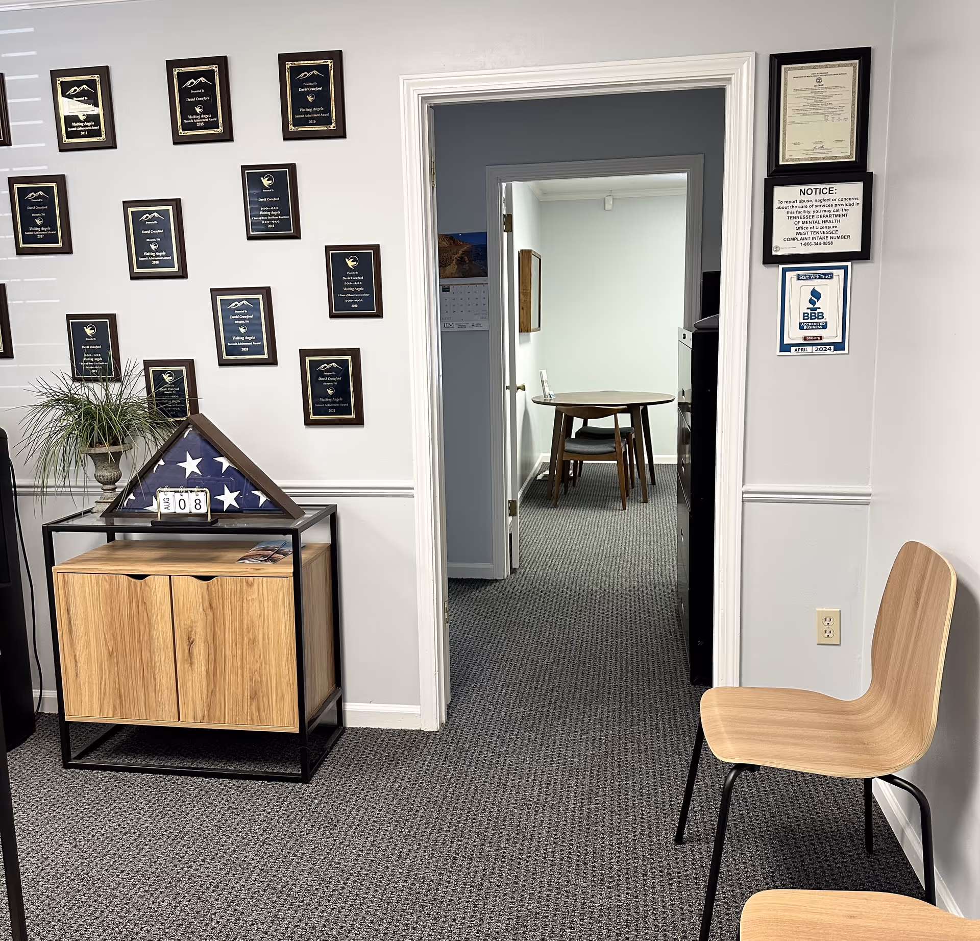 Interior hallway of a senior facility featuring wall plaques, a cabinet, and nearby chairs.