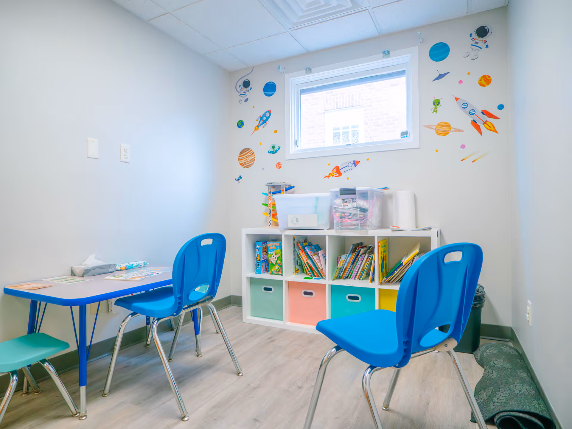 Bright children's playroom with blue plastic chairs, bookshelves, and space-themed wall decals.