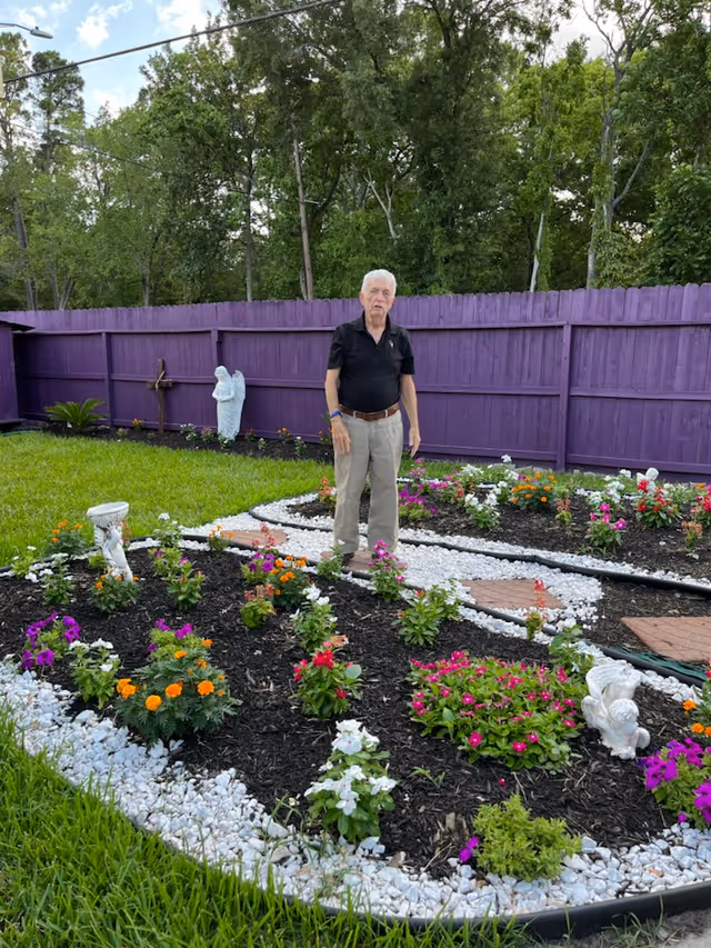 Older man stands in a landscaped garden with colorful flowers and a purple fence.