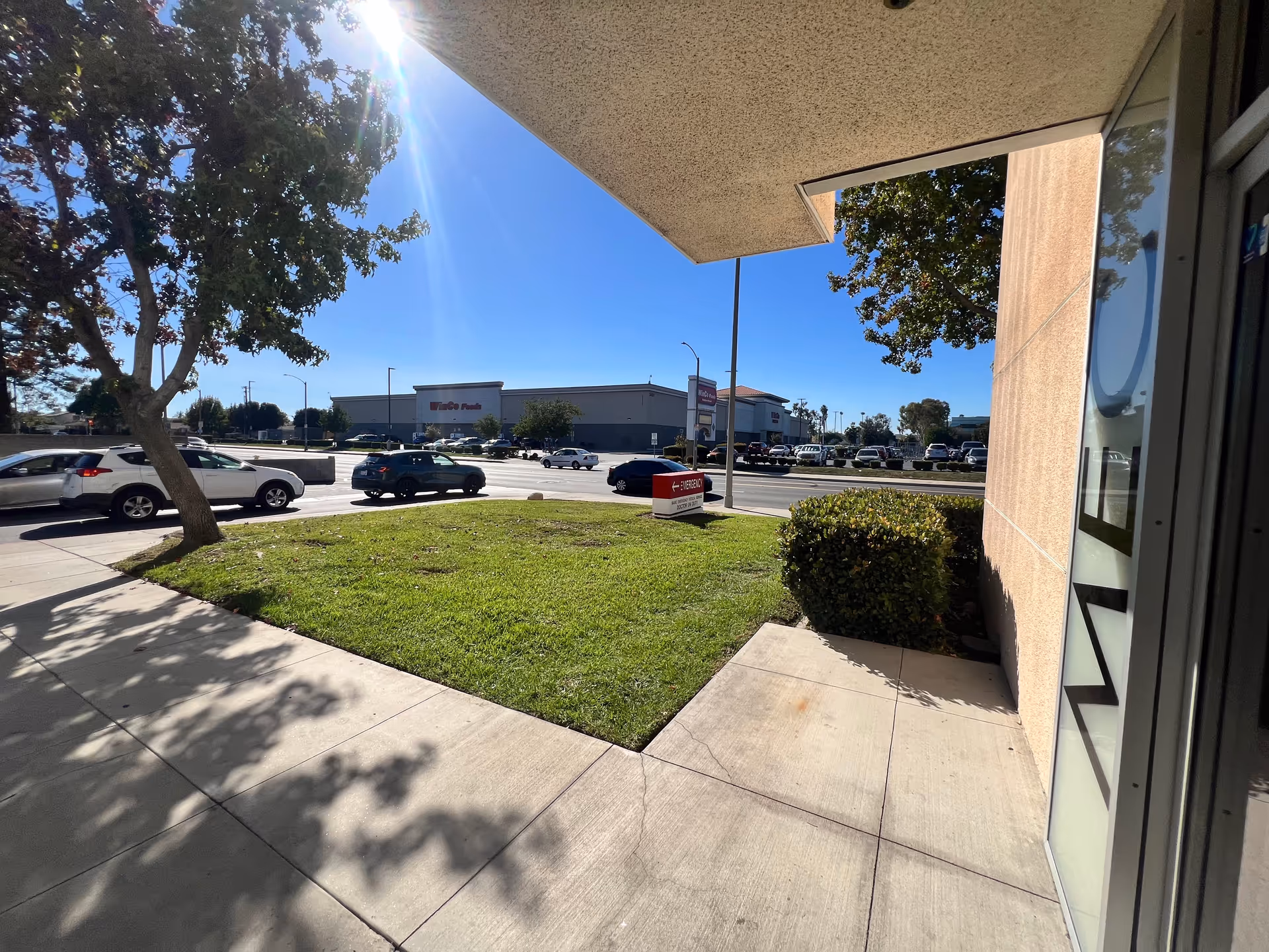Exterior view of a beige building entrance with sidewalk, grass, trees, and a parking lot.