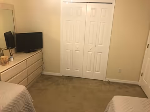 Two beds flank a dresser and TV in a small beige bedroom with closet doors.