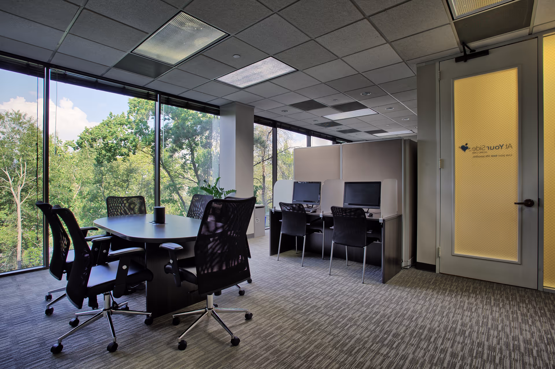 Modern office space with a round conference table, chairs, and cubicles beside large windows.