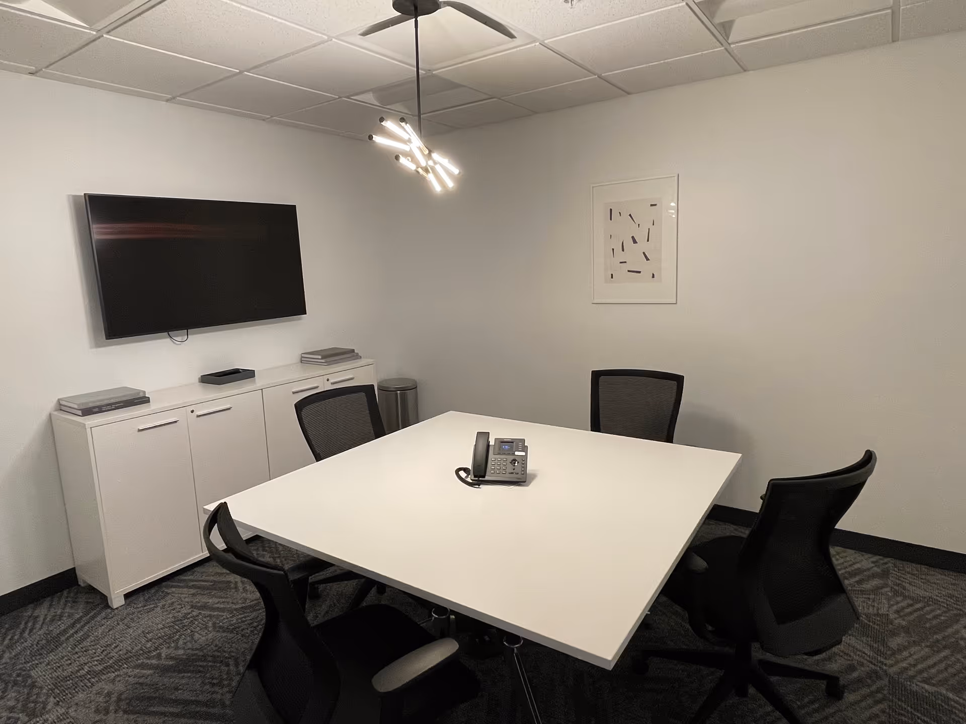A small meeting room with a white table, black chairs, wall-mounted TV, and cabinets.