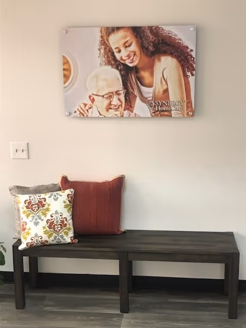 A dark wooden bench with cushions against a wall under a SYNERGY HomeCare poster.