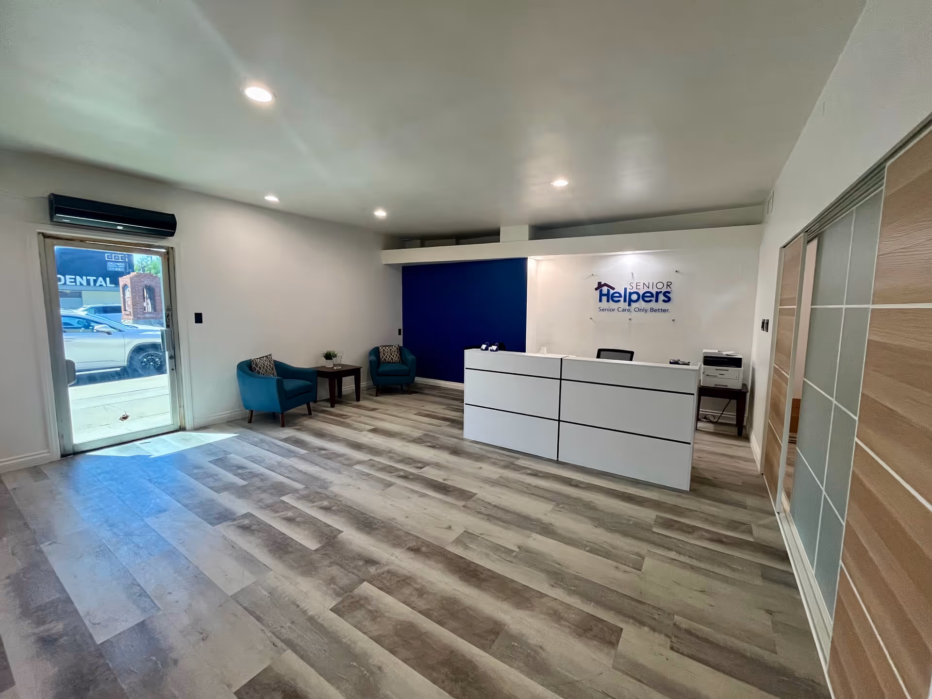 Interior reception area in Senior Helpers with a white front desk, blue accent wall, teal chairs.