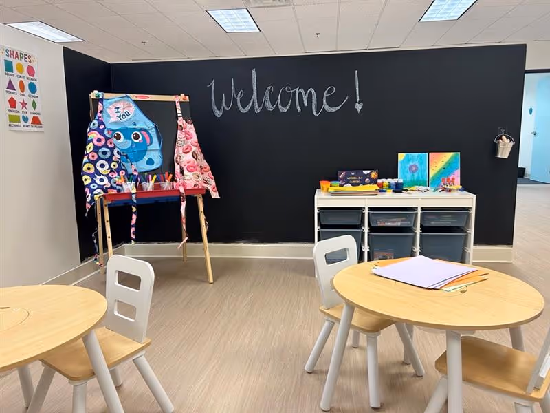 Chalkboard wall with 'Welcome', craft stations, round tables, and storage cubbies.
