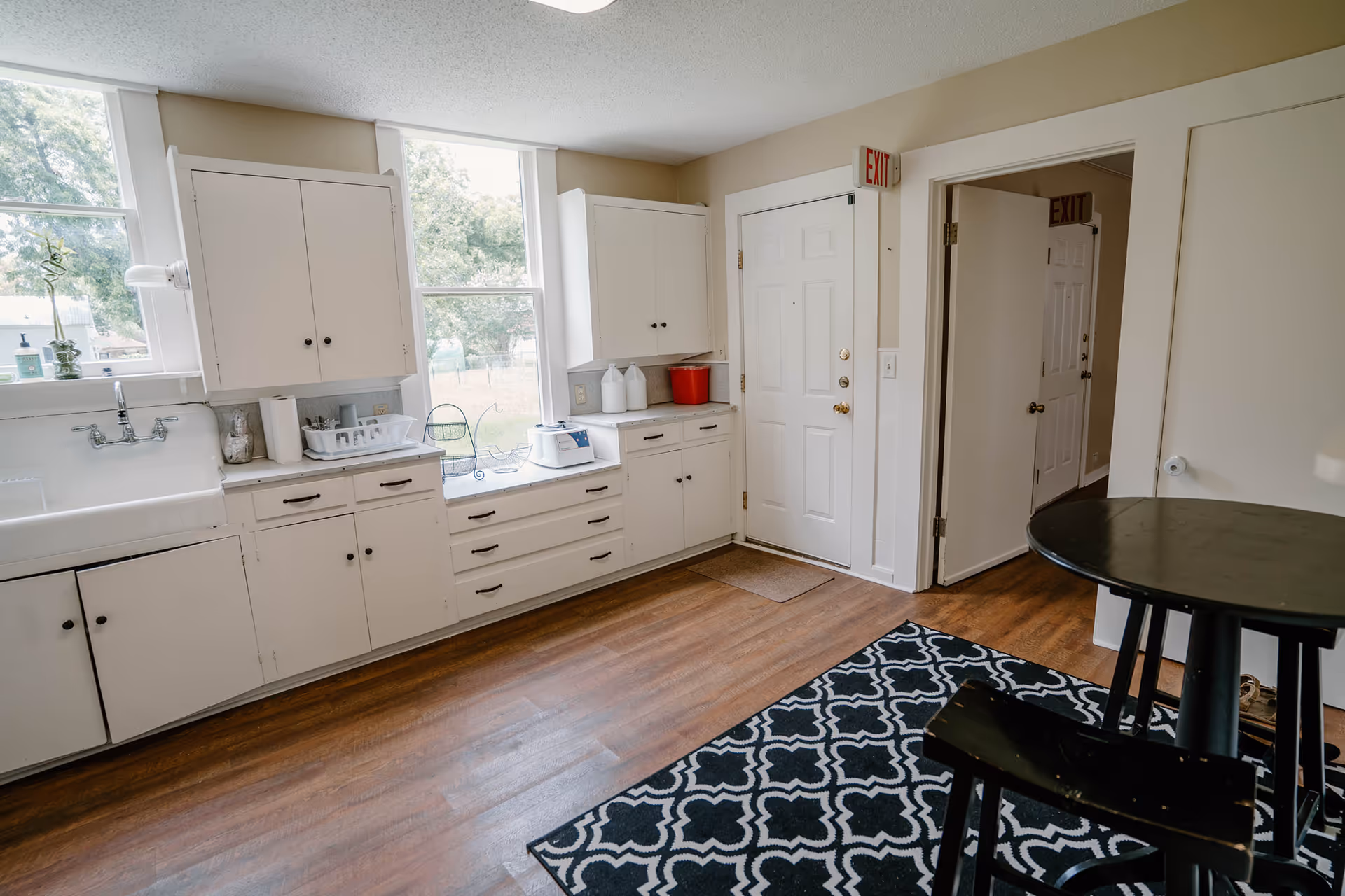 Bright white kitchen with cabinets, sink, windows, and a round black table.