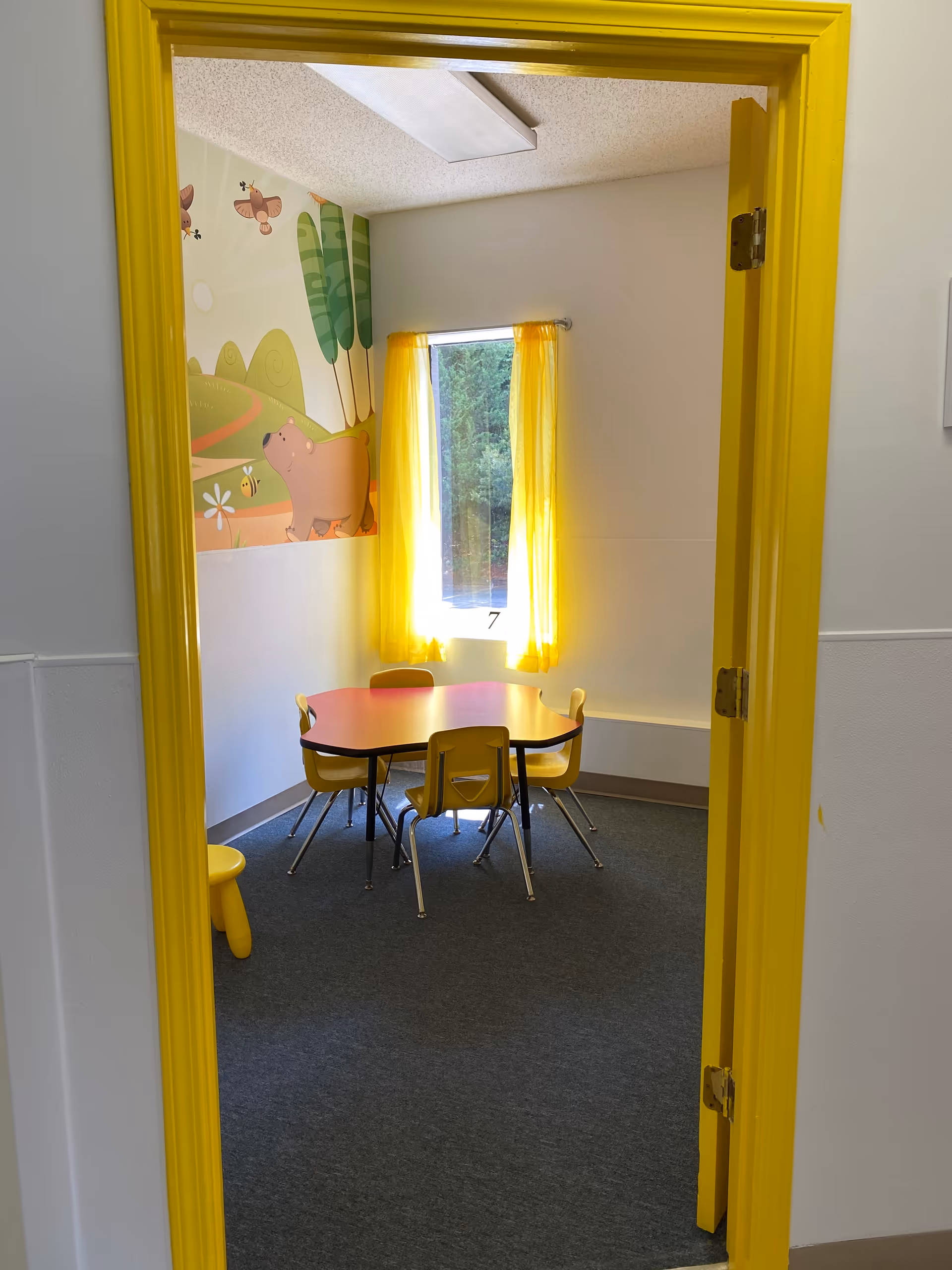 Bright yellow doorway frames a small muraled dining room with a pink table and yellow chairs.