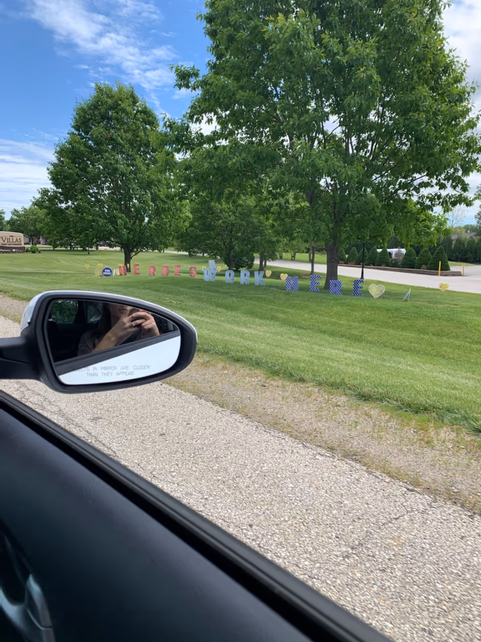 Car side mirror shows grassy exterior with colorful 'Heroes Work Here' sign along the road.