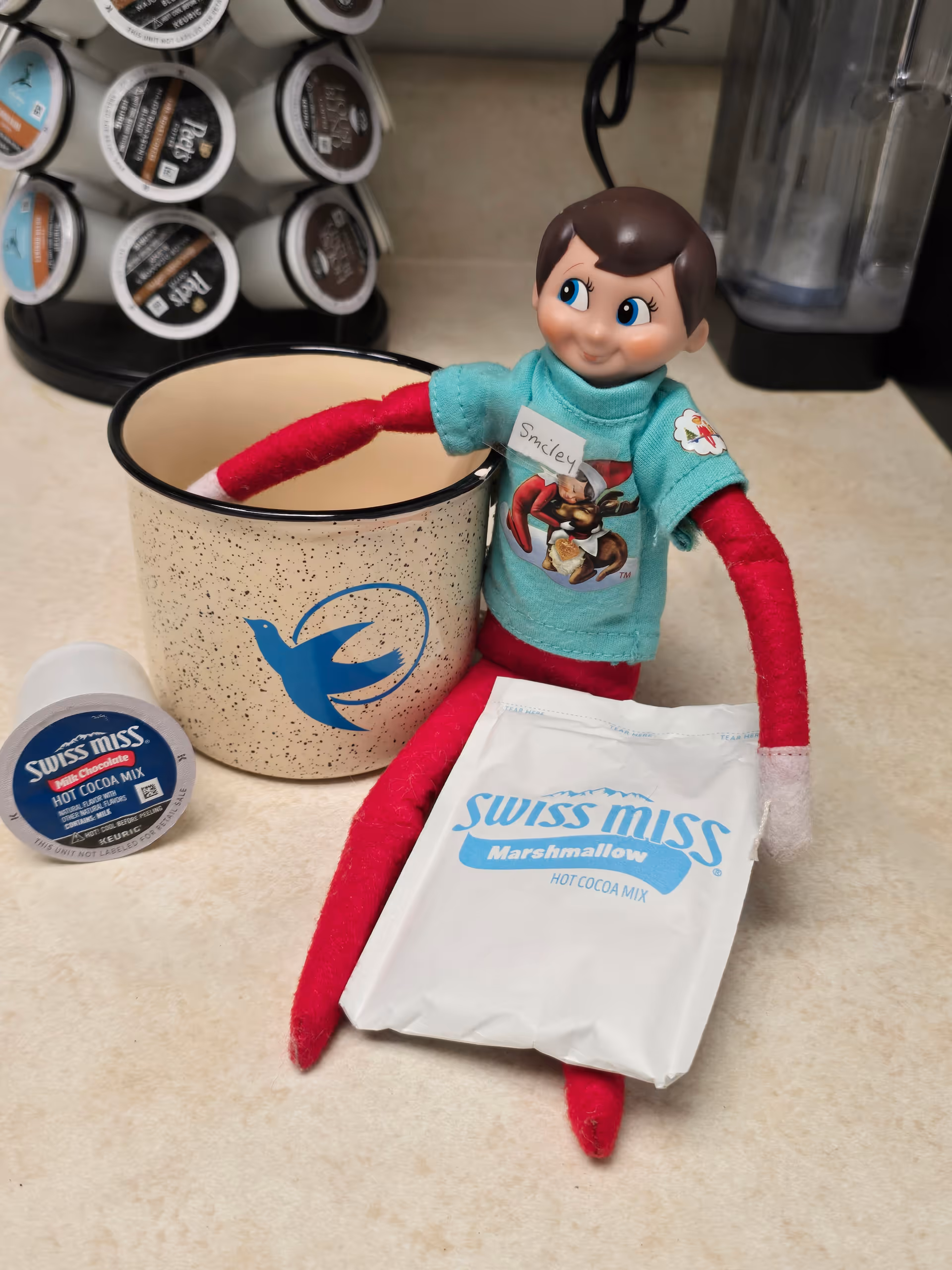 A small doll sits on a kitchen counter beside cocoa mix and a coffee pod rack.