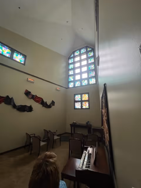 Chapel-style interior with stained glass windows, rows of chairs, a piano, and a seated person.