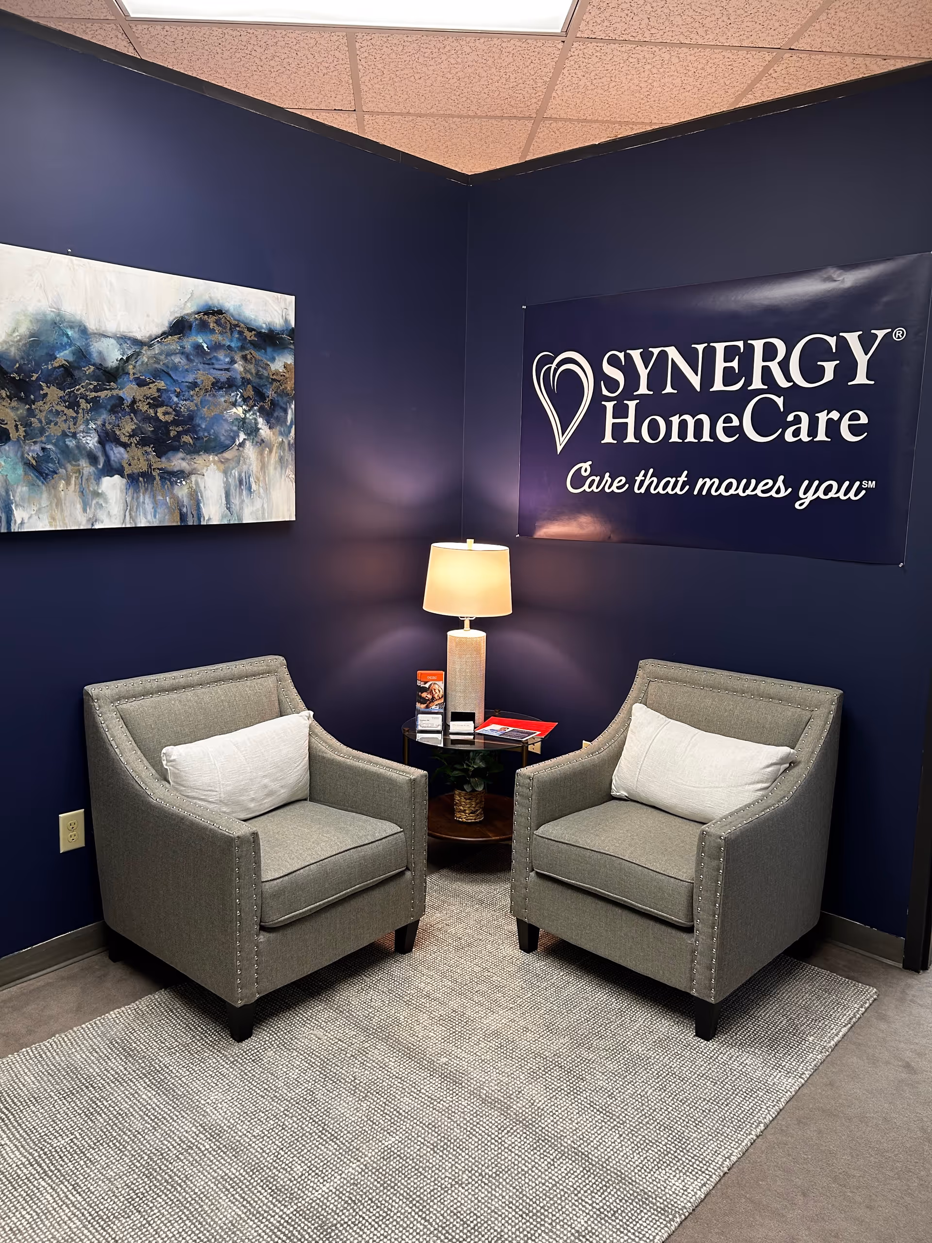 Two gray armchairs with a round table and lamp in a navy lounge with Synergy banner