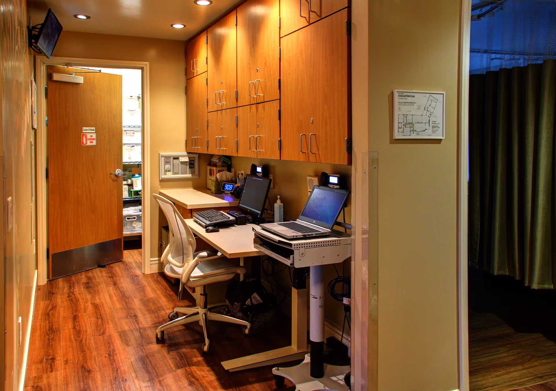 Office workstation along a hallway with laptops, desktop screen, and wooden cabinets.