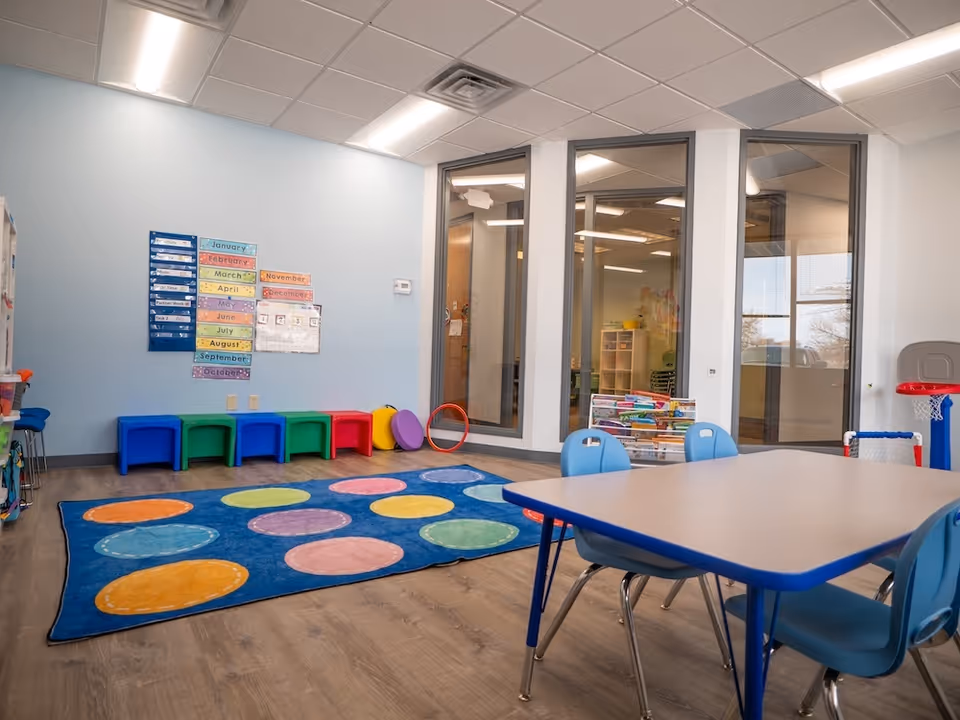 Bright pediatric therapy room with colorful rug, tables, chairs, and wall charts.