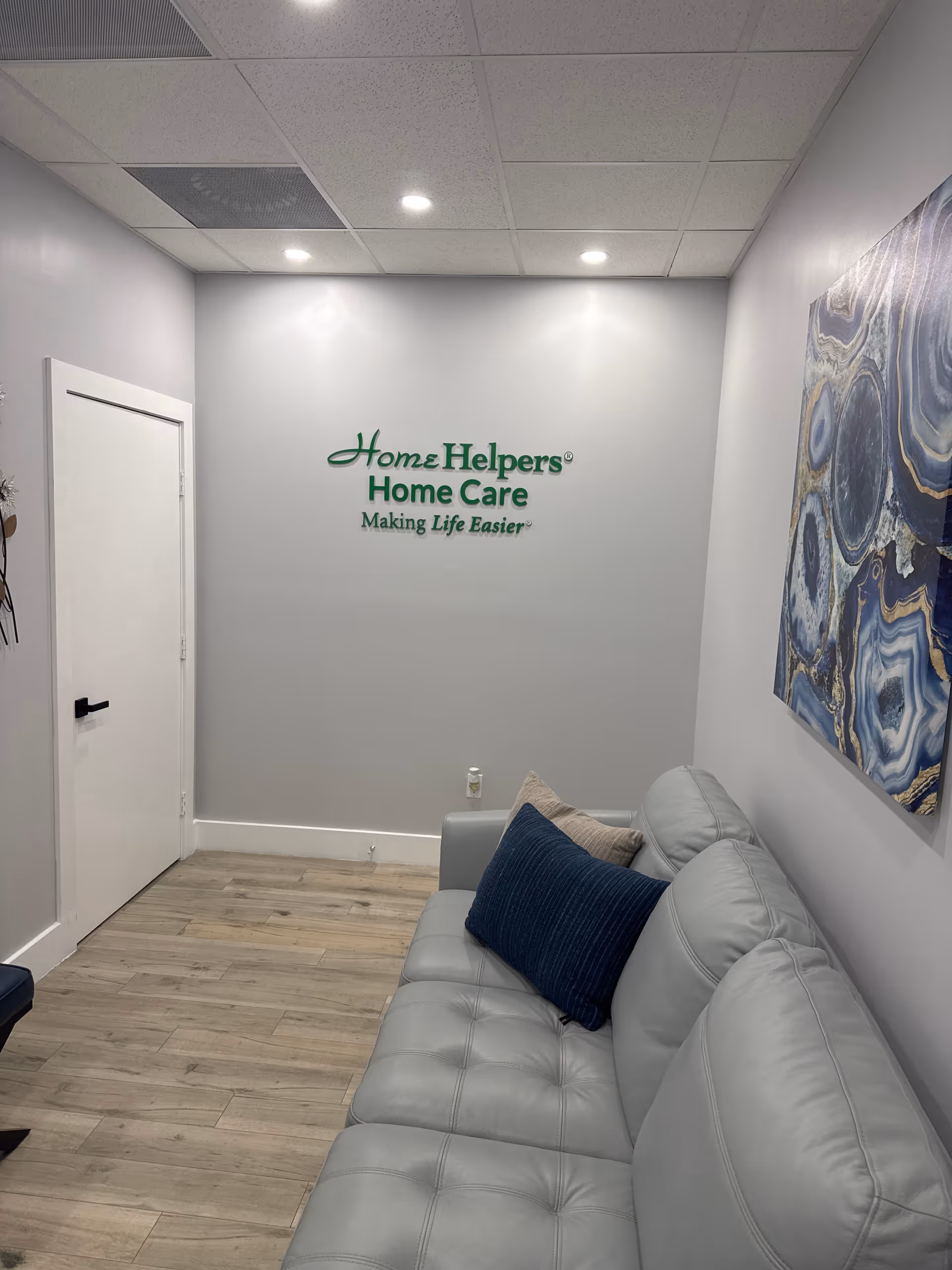 Interior waiting area at Home Helpers Home Care with a gray couch and wall logo.