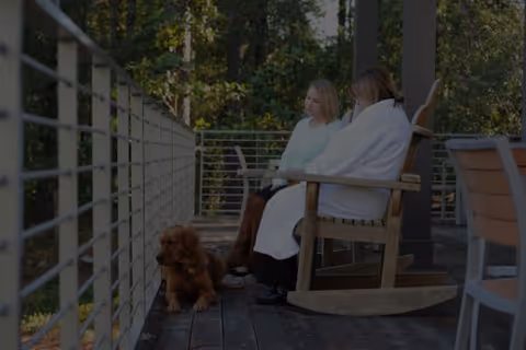 Two residents sit on a wooden outdoor porch with a small dog beside them.