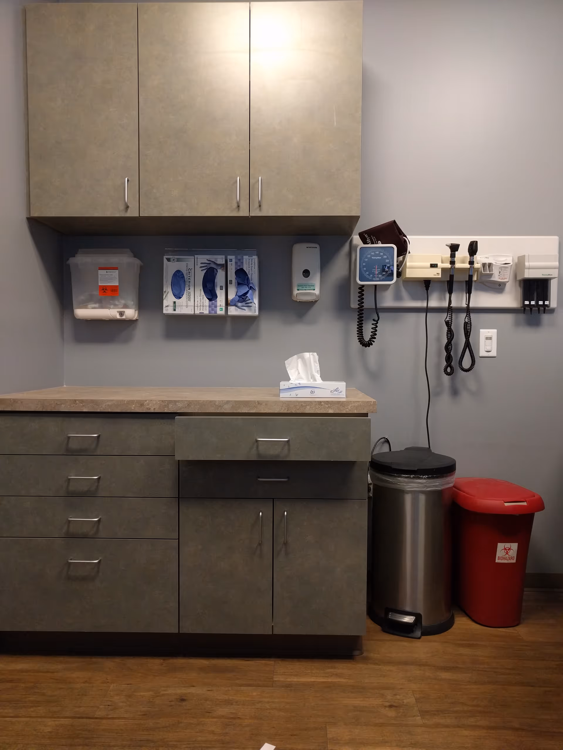 Clinic-style examination area with gray cabinets, medical devices, tissue box, and waste bins.