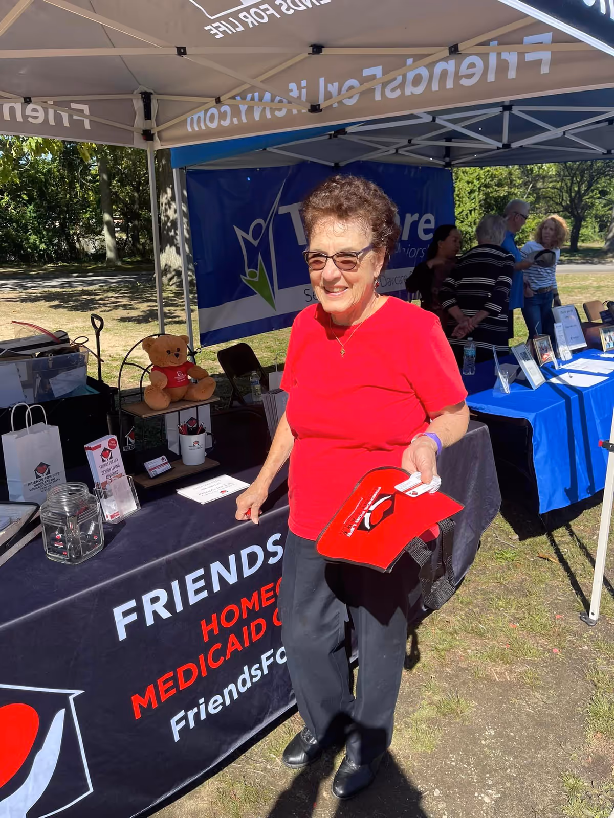 A senior woman in a red shirt stands at an outdoor booth under a canopy.