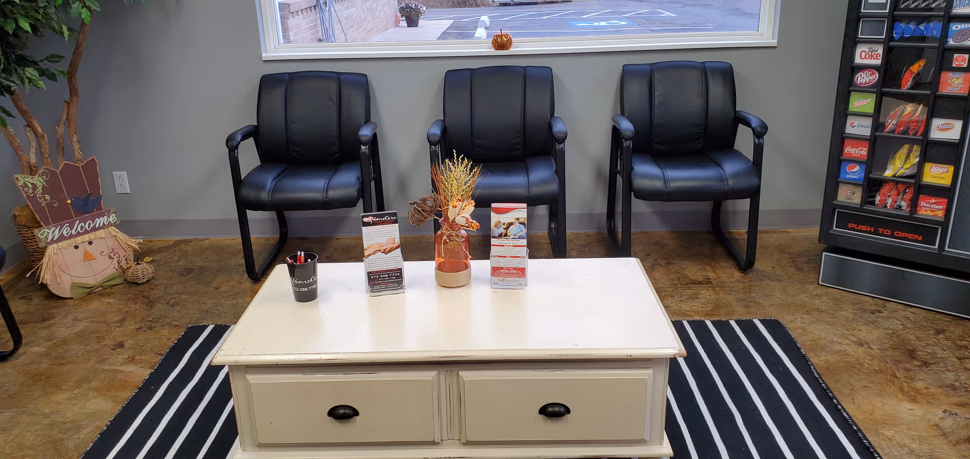 Waiting area with three black chairs, a coffee table, brochures, a vending machine, and plants.