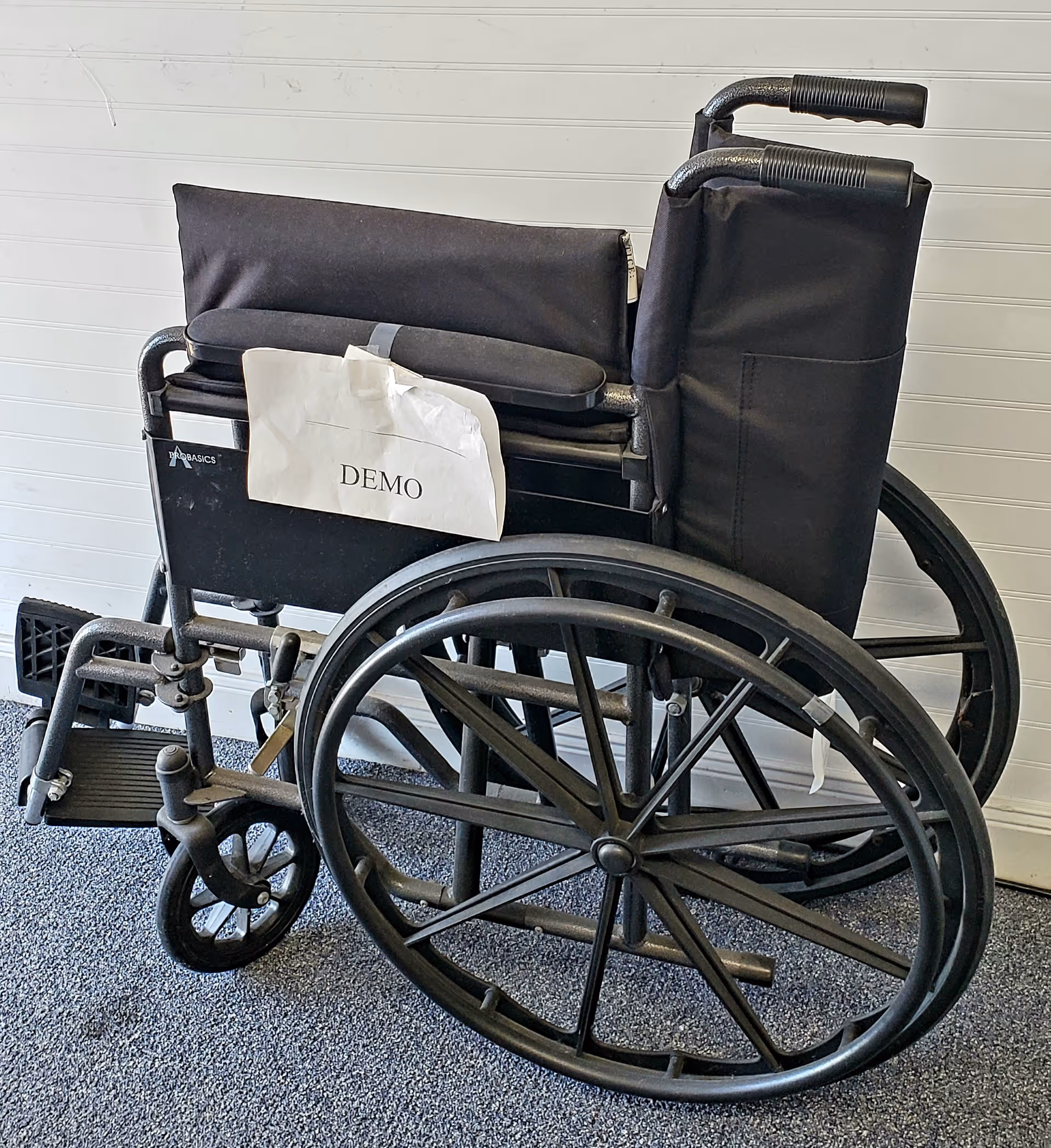 A manual wheelchair labeled DEMO sits indoors against a light-paneled wall.