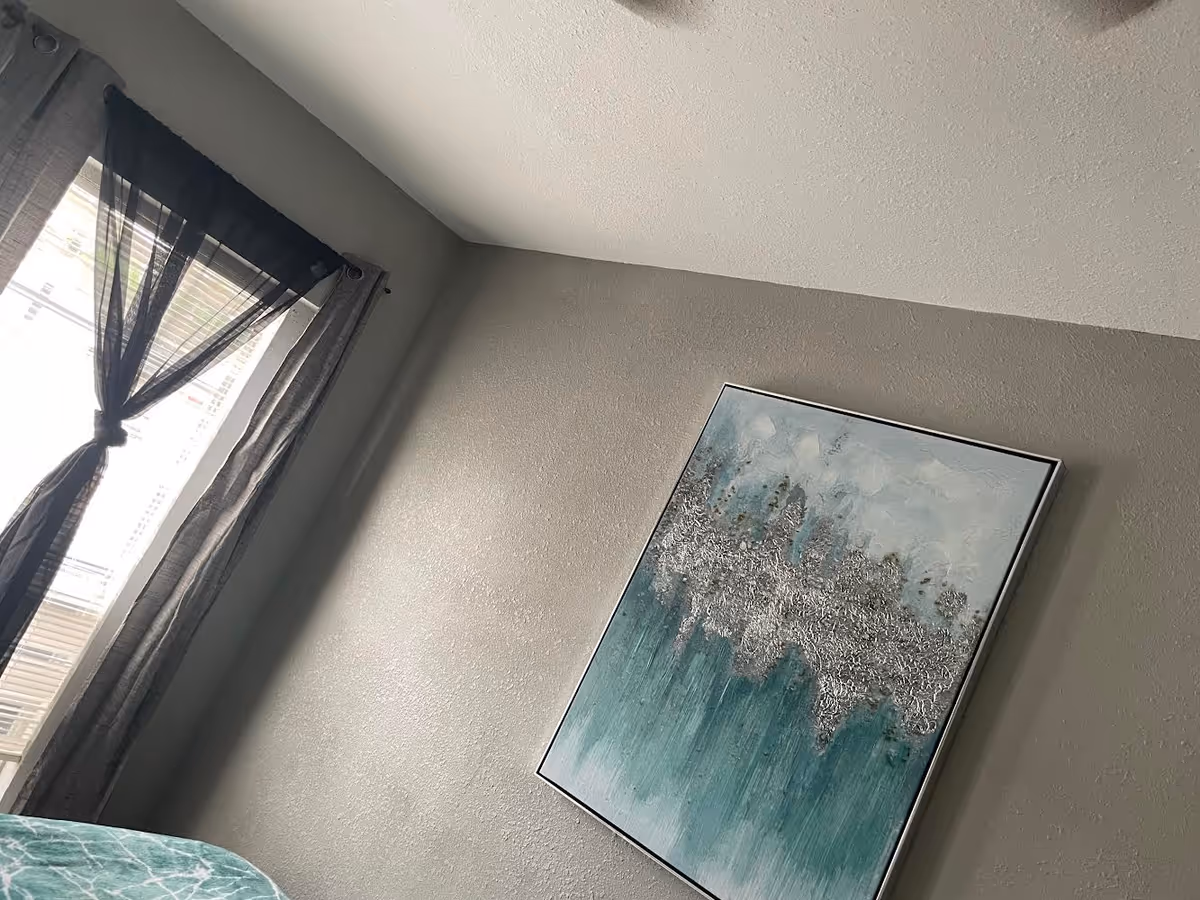 Corner of a bedroom with a window, sheer curtains, and abstract teal wall art.