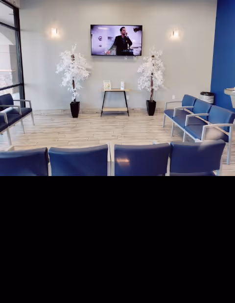 Waiting area with blue chairs, a wall-mounted TV, and white decorative trees.