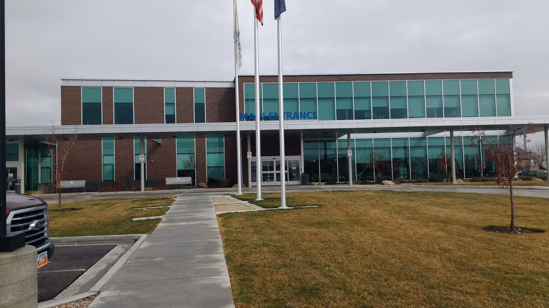 Brick building with glass facade and a main entrance, flagpoles, and a sidewalk.