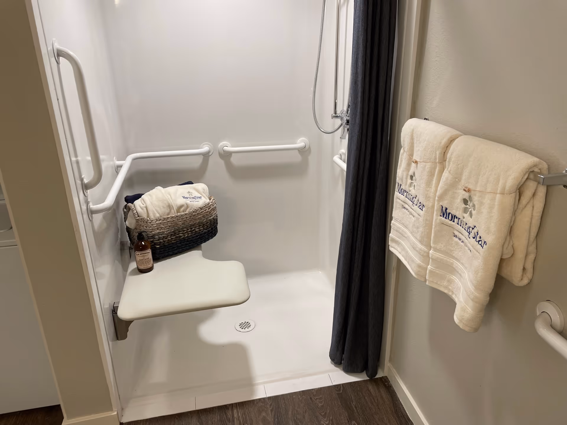 Accessible bathroom shower with a chair, grab bars, towel rack, and curtain.