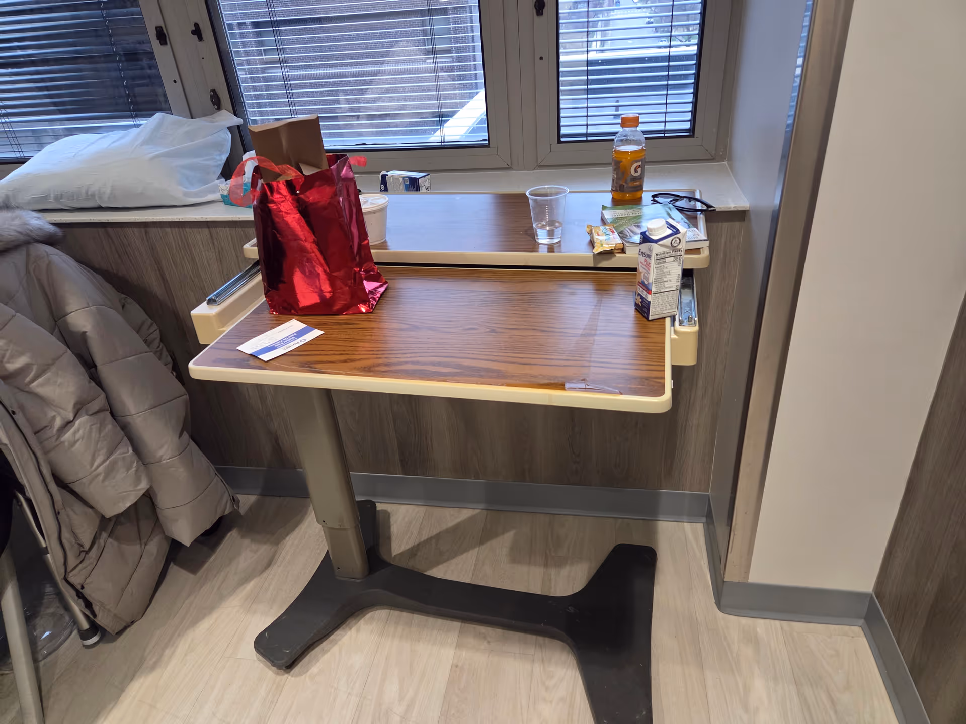 Hospital room desk with snacks, drink, and personal items by window.