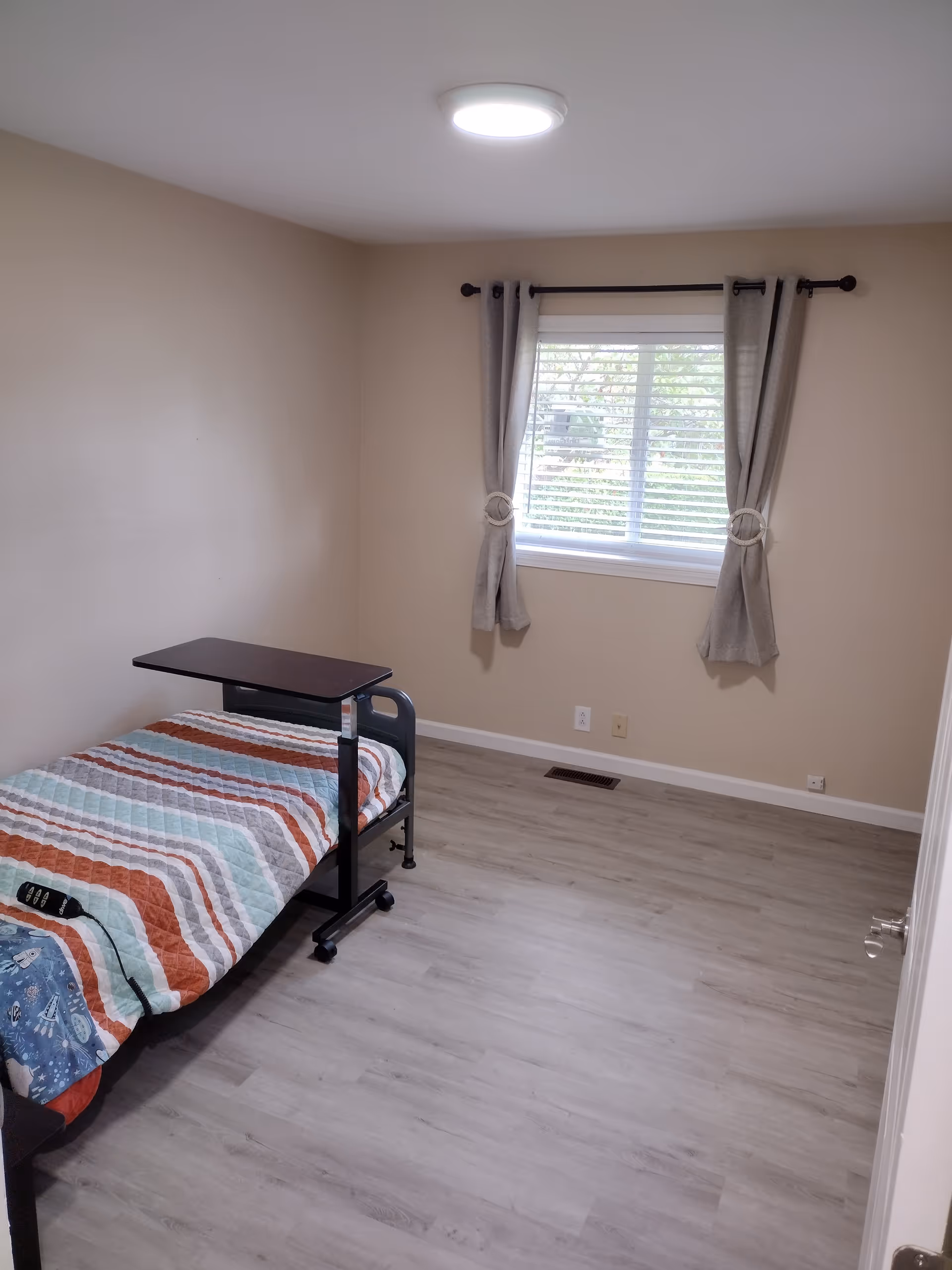 Small beige bedroom with a single bed, overbed table, and window with curtains.